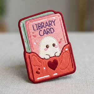 May include: An embroidered patch featuring a pink library card with the text LIBRARY CARD in purple. A small white ghost with pink cheeks peeks out from a red pocket at the bottom, which is decorated with a dark red heart and abstract shapes.