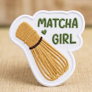 May include: A white embroidered patch featuring a golden-brown bamboo matcha whisk with a dark brown band. The text MATCHA GIRL is stitched in dark green thread above a small green heart, all set against a white background with a white border.