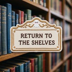 May include: A rectangular embroidered patch with a cream-colored background and a gold border. The text "RETURN TO THE SHELVES" is stitched in dark brown. A small white ghost with a smiling face is centered above the text.