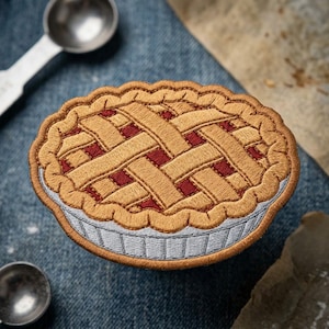 May include: An embroidered patch shaped like a round fruit pie with a golden-brown lattice crust and visible red filling. The pie features a scalloped edge and a light blue fluted base, rendered in detailed thread work with a textured appearance.