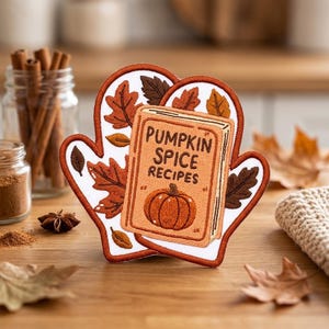 May include: An embroidered patch shaped like an oven mitt featuring a white background with brown and orange autumn leaves. Centered on the mitt is a rectangular book illustration with the text PUMPKIN SPICE RECIPES and a small pumpkin graphic.