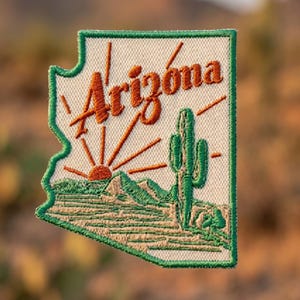 May include: An embroidered patch shaped like the state of Arizona with a cream-colored background and a green border. It features a desert landscape with a green saguaro cactus, tan terrain, and a rising orange sun with the word Arizona in orange.