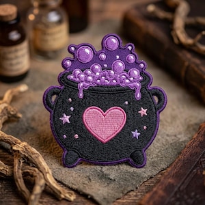 May include: An embroidered patch featuring a black cauldron with a pink heart at the center. The cauldron is filled with bubbling purple liquid that overflows the rim. Small embroidered stars in light pink and purple surround the heart.
