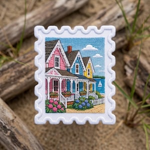 May include: A rectangular embroidered patch shaped like a postage stamp featuring a row of three colorful houses in pink, blue, and yellow. The scene includes a blue sky with white clouds, a body of water with a sailboat, and blooming flowers.