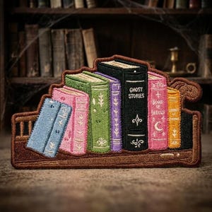May include: An embroidered patch featuring a row of colorful books on a brown shelf. The books are blue, pink, green, purple, black with the text GHOST STORIES, pink with the text BOOK OF SPELLS and a crescent moon, and yellow with the text BOOK OF SHADOWS.