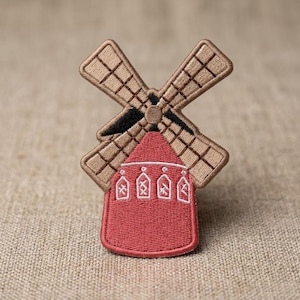 May include: An embroidered patch shaped like a windmill with a red base and tan blades. The red base features four white outlined windows with cross-stitch details. The windmill blades are tan with black accents at the center hub.