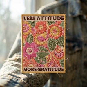 May include: A rectangular embroidered patch featuring a floral pattern in shades of pink, orange, yellow, and green. The text LESS ATTITUDE is stitched in dark brown at the top, and MORE GRATITUDE is stitched in dark brown at the bottom.