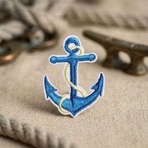 May include: An embroidered patch featuring a blue anchor with a white border and a yellow rope wrapped around the central shaft. The design is rendered in detailed thread work, showcasing a nautical motif with clear, defined stitching throughout.