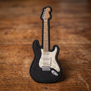 May include: An embroidered patch shaped like an electric guitar with a black body, a white pickguard, and a tan neck. The design features detailed stitching for the strings, frets, and tuning pegs, all outlined with a thick black border.