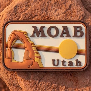 May include: A rectangular Moab, Utah souvenir patch. The patch features a brown border, a cream-colored background, and the words "MOAB" and "Utah" in brown. It also includes an archway and a yellow circle.