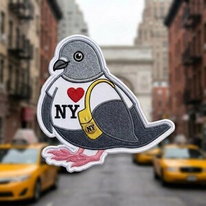 May include: An embroidered patch featuring a gray pigeon wearing a white t-shirt with a red heart, the text NY, and a yellow shoulder bag with an NY logo. The pigeon has pink feet and is outlined in white against a blurred city street background.