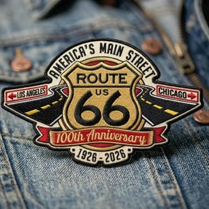 May include: An embroidered patch featuring a gold Route 66 shield with black text, flanked by black roads pointing to Los Angeles and Chicago. It includes the text America's Main Street, 100th Anniversary, and 1926-2026 in black and red on a cream background.