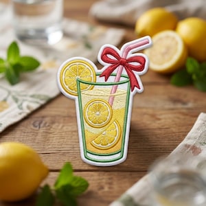 May include: An embroidered patch shaped like a glass of lemonade with yellow liquid, two lemon slices inside, a pink straw, and a red bow tied around the straw. The patch features a white border and detailed stitching in yellow, green, pink, and red.