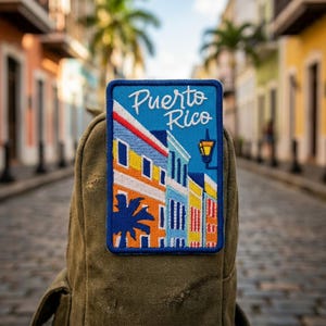 May include: A rectangular embroidered patch featuring the text Puerto Rico in white script. The design depicts colorful buildings in orange, yellow, and blue, a black palm tree silhouette, and a black street lamp against a blue sky background.