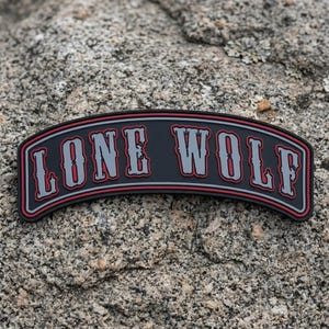 May include: A black, curved patch with the words "LONE WOLF" in gray, outlined in red and white. The patch is set against a textured, gray rock background. The text is in a bold, stylized font.