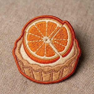 May include: A circular embroidered patch featuring a detailed orange slice resting atop a tan tart base. The orange slice displays vibrant orange segments with yellow highlights, while the tart crust is rendered in shades of light brown.