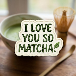 May include: An embroidered patch with a cream-colored background and a scalloped edge. The patch features dark green text that reads I LOVE YOU SO MATCHA! with a small green leaf icon at the end of the text, designed for fabric application.