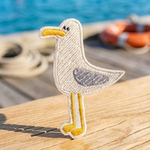 May include: An embroidered patch shaped like a seagull. The bird has a cream-colored body, a bright yellow beak and feet, and a grey wing detail. The design features simple black stitching for the eye, with a clean white border outline.