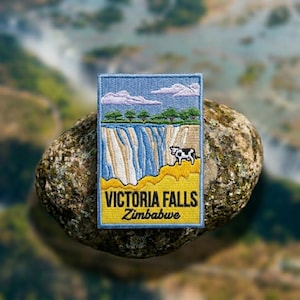 May include: A rectangular embroidered patch featuring a landscape of Victoria Falls in Zimbabwe. The design includes a blue sky with clouds, green trees, a waterfall, and a black and white cow on a yellow foreground with the text Victoria Falls Zimbabwe.
