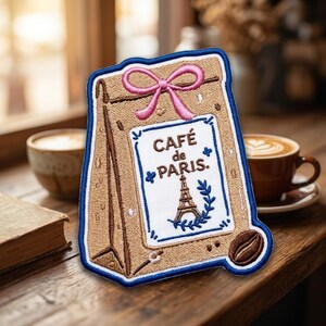May include: An embroidered patch shaped like a tan paper bag with a pink bow at the top. The center features a white label with blue text reading CAFÉ de PARIS, a small Eiffel Tower illustration, and a single brown coffee bean at the bottom right.