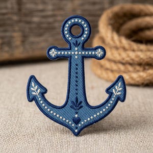 May include: A blue anchor-shaped embroidered patch featuring a dark blue border and intricate white stitching details. The design includes small white dots, floral motifs, and a small heart shape at the base, all set against a textured denim-like fabric.