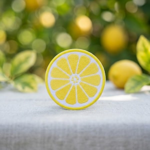 May include: A circular embroidered patch featuring a stylized lemon slice design. The patch displays a vibrant yellow center with distinct segments separated by white lines, all enclosed within a bright yellow circular border edge.
