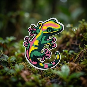 May include: An embroidered patch shaped like a gecko, featuring a vibrant color palette of dark green, bright yellow, and pink. The lizard design has a curved tail and detailed stitching, outlined in white, showcasing a stylized reptile form.