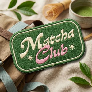 May include: A rectangular embroidered patch with rounded corners featuring a dark green background. The text "Matcha" is stitched in cream, while "Club" is in pink. Two small cream-colored starburst symbols flank the text on the patch.