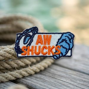 May include: A rectangular embroidered patch featuring a light blue background with dark blue oyster shell illustrations on the left and right sides. The text AW SHUCKS is stitched in bold orange capital letters across the center of the patch.