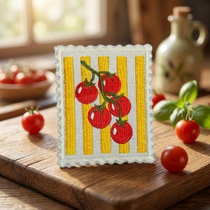May include: A rectangular embroidered patch with a scalloped edge featuring a design of red tomatoes on a green vine against a background of vertical yellow and white stripes. The patch is positioned upright on a wooden surface.