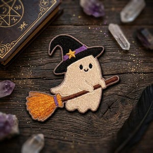 May include: An embroidered patch featuring a light beige ghost wearing a black witch hat with a purple band and a yellow star. The ghost is riding a brown broomstick with orange bristles. The patch has a dark brown stitched border.