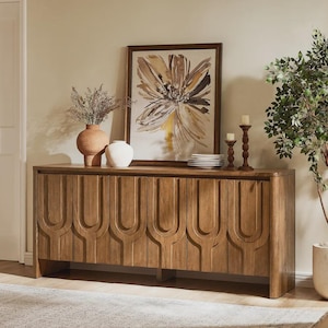 May include: A wooden sideboard with a carved geometric pattern on the doors. It's topped with a framed floral painting, vases, candles, and plates. The colour scheme is neutral with warm wood tones, typical of modern furniture.