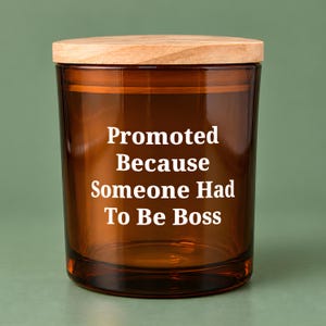 May include: A cylindrical amber glass candle jar with a light-colored wooden lid. The front of the glass features white text that reads: Promoted Because Someone Had To Be Boss. The container has a smooth, transparent finish.