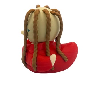 Peut inclure: Une figurine de canard en caoutchouc avec une t&ecirc;te beige p&acirc;le et de longues m&egrave;ches de cheveux brunes torsad&eacute;es en forme de corde. Le canard porte une veste rouge vif avec des d&eacute;tails de col et une petite aile beige p&acirc;le visible sur le c&ocirc;t&eacute;.