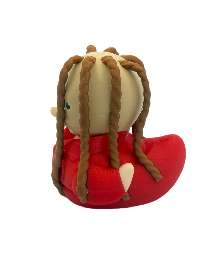 Peut inclure: Une figurine de canard en caoutchouc avec une t&ecirc;te beige p&acirc;le et de longues m&egrave;ches de cheveux brunes torsad&eacute;es en forme de corde. Le canard porte une veste rouge vif avec des d&eacute;tails de col et une petite aile beige p&acirc;le visible sur le c&ocirc;t&eacute;.