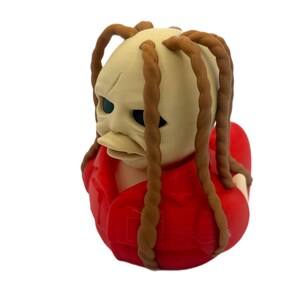 Peut inclure: Une figurine de canard en caoutchouc avec une t&ecirc;te p&acirc;le et textur&eacute;e, dot&eacute;e de longues m&egrave;ches de cheveux tress&eacute;es brunes. Le personnage porte une chemise rouge vif &agrave; col et pr&eacute;sente des orbites sombres et un bec plat de canard.