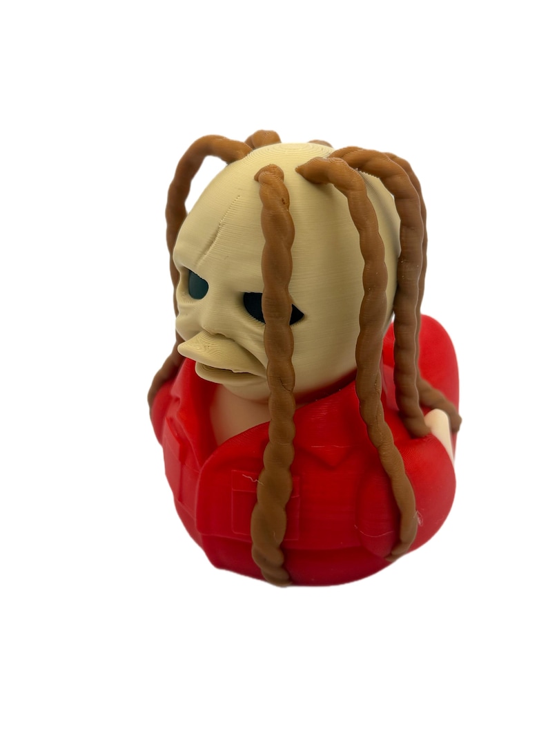 Peut inclure: Une figurine de canard en caoutchouc avec une t&ecirc;te p&acirc;le et textur&eacute;e, dot&eacute;e de longues m&egrave;ches de cheveux tress&eacute;es brunes. Le personnage porte une chemise rouge vif &agrave; col et pr&eacute;sente des orbites sombres et un bec plat de canard.