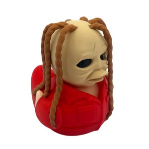 Peut inclure: Une figurine de canard en caoutchouc repr&eacute;sentant une personne avec une t&ecirc;te p&acirc;le et lisse, des yeux sombres et de longs cheveux bruns tress&eacute;s. La figurine porte une chemise rouge vif &agrave; col avec une petite poche sur la poitrine.