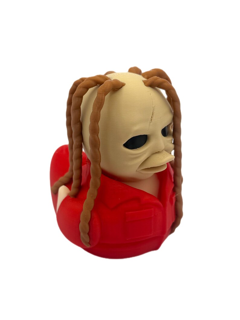 Peut inclure: Une figurine de canard en caoutchouc repr&eacute;sentant une personne avec une t&ecirc;te p&acirc;le et lisse, des yeux sombres et de longs cheveux bruns tress&eacute;s. La figurine porte une chemise rouge vif &agrave; col avec une petite poche sur la poitrine.