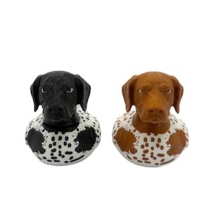 May include: Two dog-shaped bottle stoppers. One is black with a white and black spotted base, the other is brown with a white and brown spotted base. The stoppers are designed to resemble the heads of dogs.