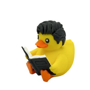 May include: A yellow rubber duck figurine with a textured black hairstyle, an orange beak, and orange feet. The duck is holding an open black book with white pages in its wings, appearing to read the text printed on the open pages of the book.
