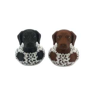 May include: Two dog-shaped figurines, one with a black head and the other with a brown head. Both figures feature white bodies covered in dark spots, floppy ears, and detailed facial features including eyes and noses, presented as a pair.