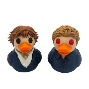 May include: Two novelty rubber ducks, each with a unique design. One duck has brown hair and a dark blue jacket, the other has blonde hair and a dark blue shirt. Both have orange beaks and black or red eyes.