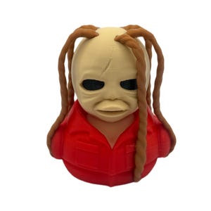 Peut inclure: Une figurine imprim&eacute;e en 3D pr&eacute;sentant une t&ecirc;te au teint p&acirc;le et lisse, dot&eacute;e d'yeux sombres et creux, avec de longues m&egrave;ches de cheveux bruns tress&eacute;s tombant sur les c&ocirc;t&eacute;s. Le personnage porte une chemise rouge vif &agrave; col avec des poches.