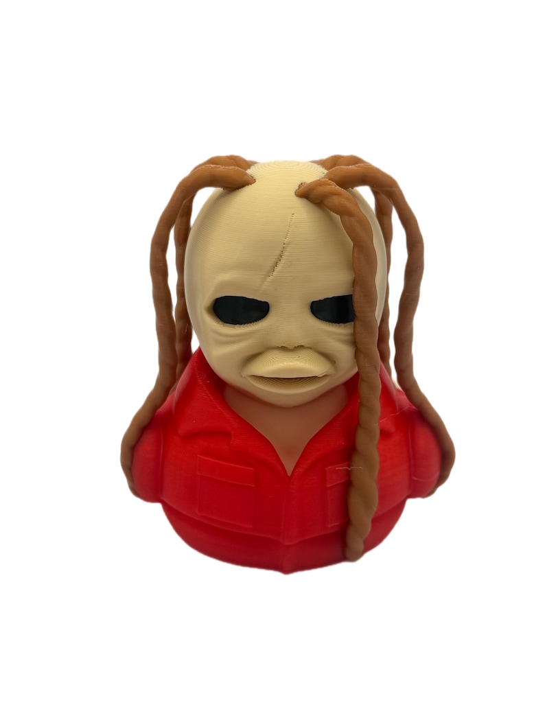 Peut inclure: Une figurine imprim&eacute;e en 3D pr&eacute;sentant une t&ecirc;te au teint p&acirc;le et lisse, dot&eacute;e d'yeux sombres et creux, avec de longues m&egrave;ches de cheveux bruns tress&eacute;s tombant sur les c&ocirc;t&eacute;s. Le personnage porte une chemise rouge vif &agrave; col avec des poches.