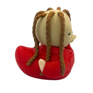 Peut inclure: Une figurine de canard en caoutchouc avec une t&ecirc;te beige p&acirc;le et de longues m&egrave;ches de cheveux tress&eacute;es brunes qui pendent. Le canard porte une veste rouge vif avec un col et une poche visibles. La figurine a une texture lisse et un bec arrondi.