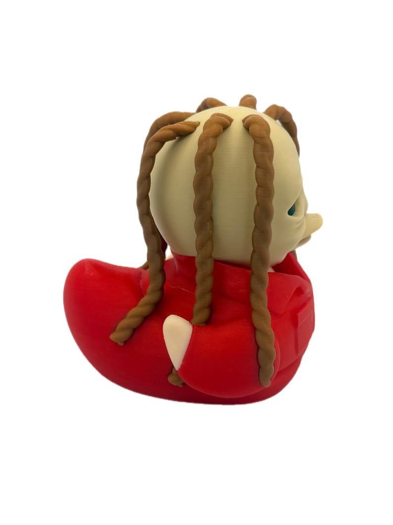 Peut inclure: Une figurine de canard en caoutchouc avec une t&ecirc;te beige p&acirc;le et de longues m&egrave;ches de cheveux tress&eacute;es brunes qui pendent. Le canard porte une veste rouge vif avec un col et une poche visibles. La figurine a une texture lisse et un bec arrondi.