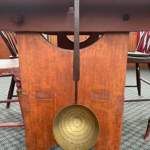 May include: A vintage brass skimmer with a long, dark metal handle. The skimmer has a circular bowl with numerous small holes arranged in concentric circles. It hangs in front of a wooden table with a warm, reddish-brown finish.