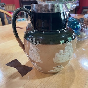 May include: A brown and green ceramic pitcher with a silver rim. The pitcher has a handle and is decorated with raised white images of a windmill, a house, a deer, and a man sitting at a table.