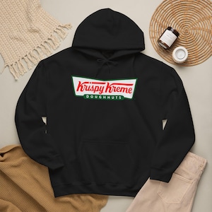 May include: A black hooded sweatshirt featuring the Krispy Kreme Doughnuts logo printed in red and green on the chest. The hoodie includes a drawstring hood, long sleeves, and a front kangaroo pocket, constructed from a solid black fabric.